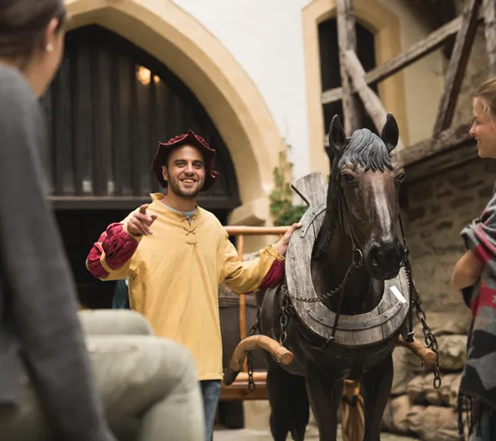 Fremdenführer in mittelalterlichem Kostüm erklärt Besuchern ein Holzpferd an historischem Ort